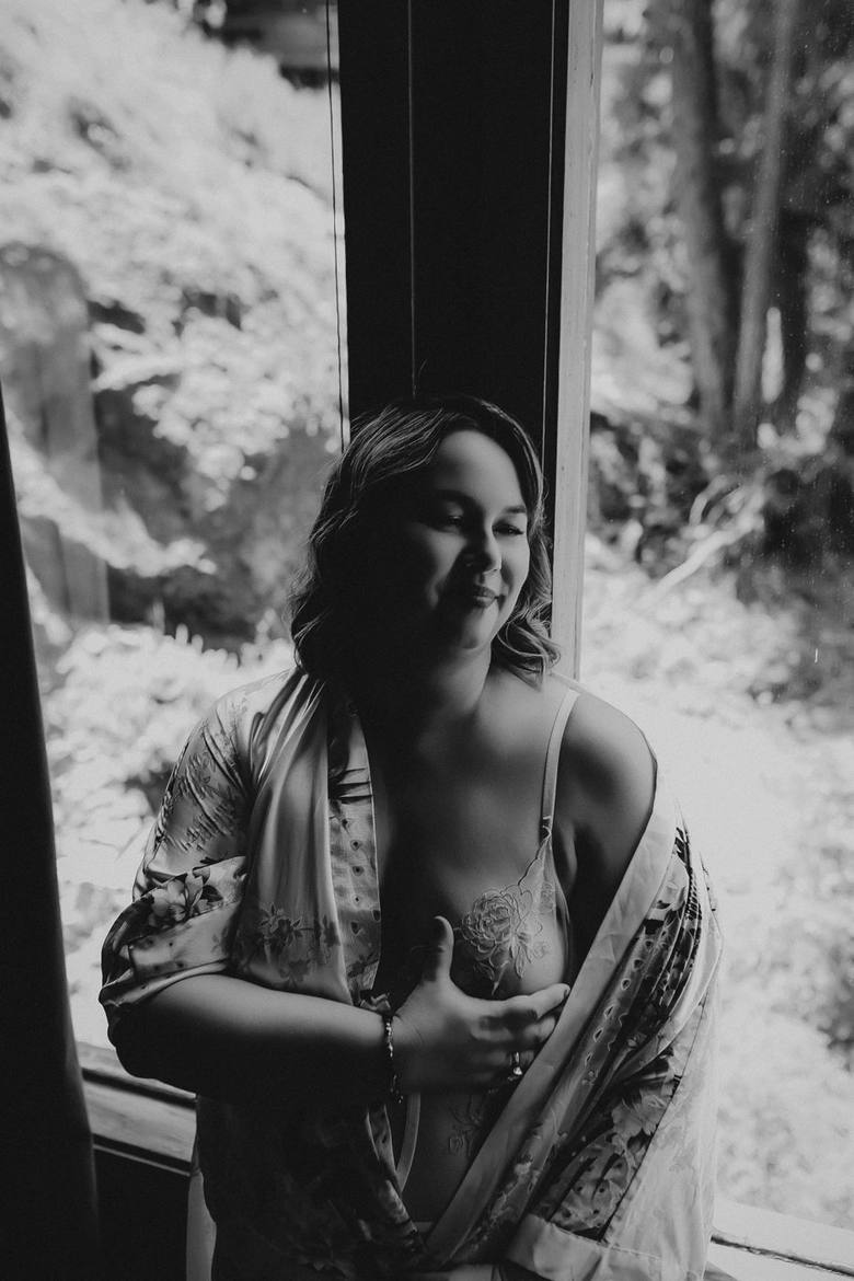 Black and white portrait of a woman in a window, robe open, hand resting on her chest