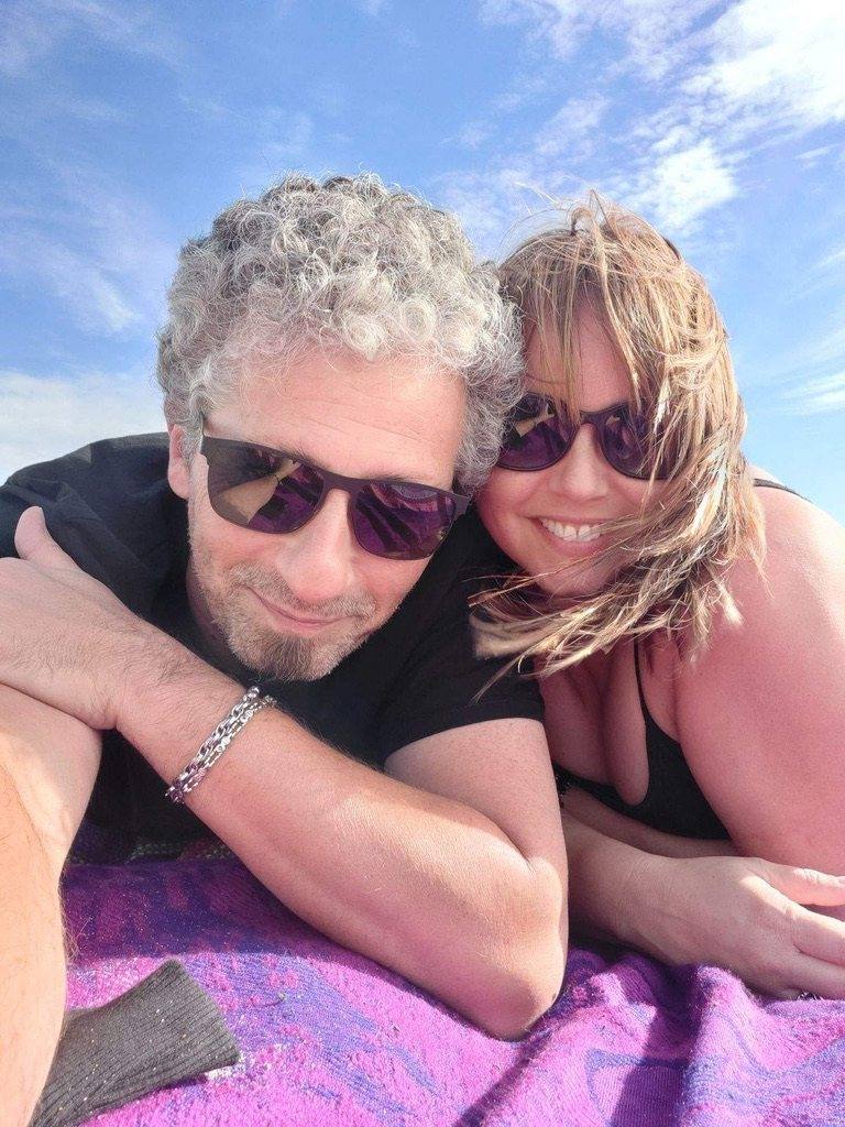 Black and white photo of a couple lying close together outdoors, both in sunglasses, her hair blowing across her smile
